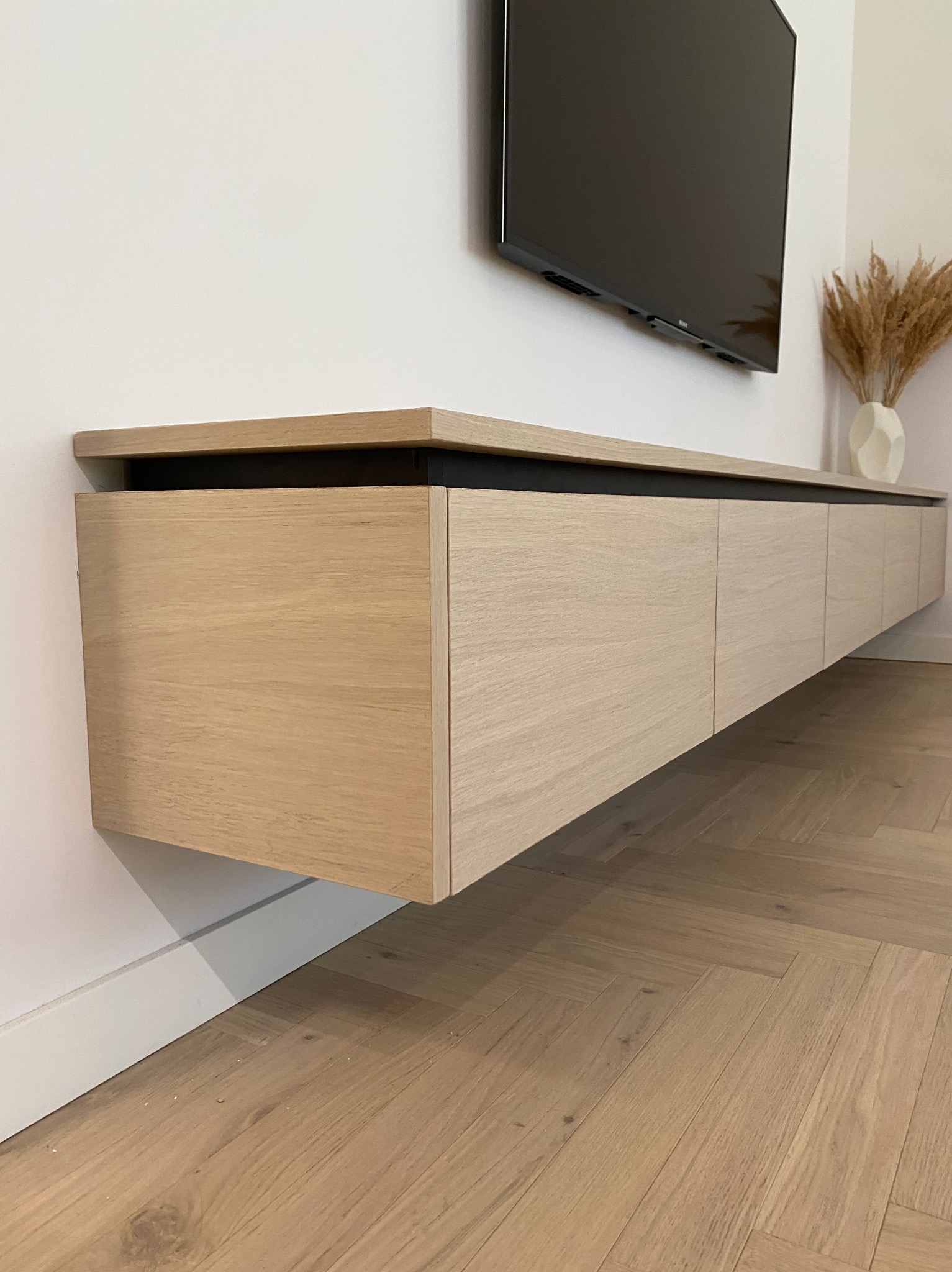 Wisząca, długa szafka RTV z jasnego drewna z ukrytymi uchwytami, zamontowana pod telewizorem na białej ścianie, z podłogą w jodełkę.