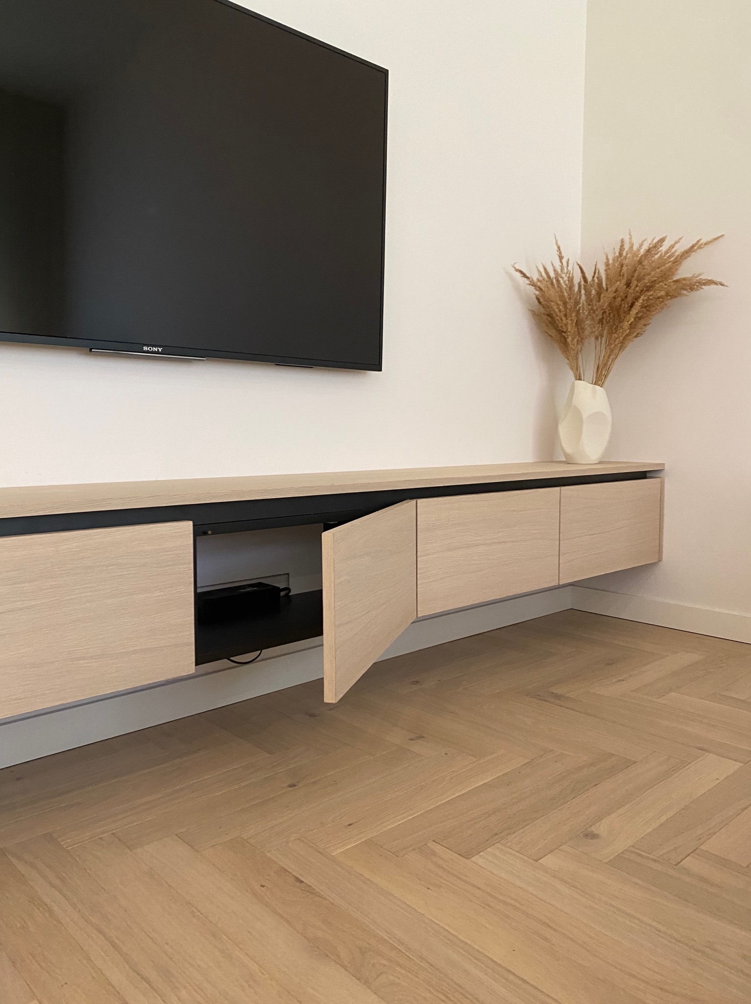 Jasna, drewniana szafka RTV wisząca na ścianie z otwartymi drzwiczkami, pod telewizorem Sony, z dekoracyjnym bukietem w wazonie na blacie i parkietem w jodełkę na podłodze.