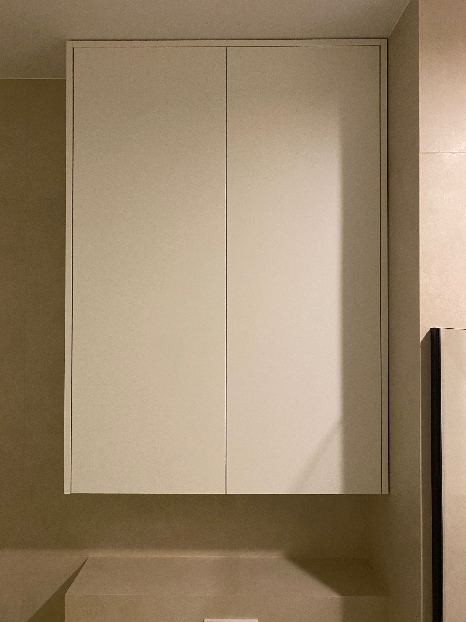 Zamknięta, kremowa szafka wisząca z dwoma drzwiczkami w nowoczesnej łazience z beżowymi płytkami.