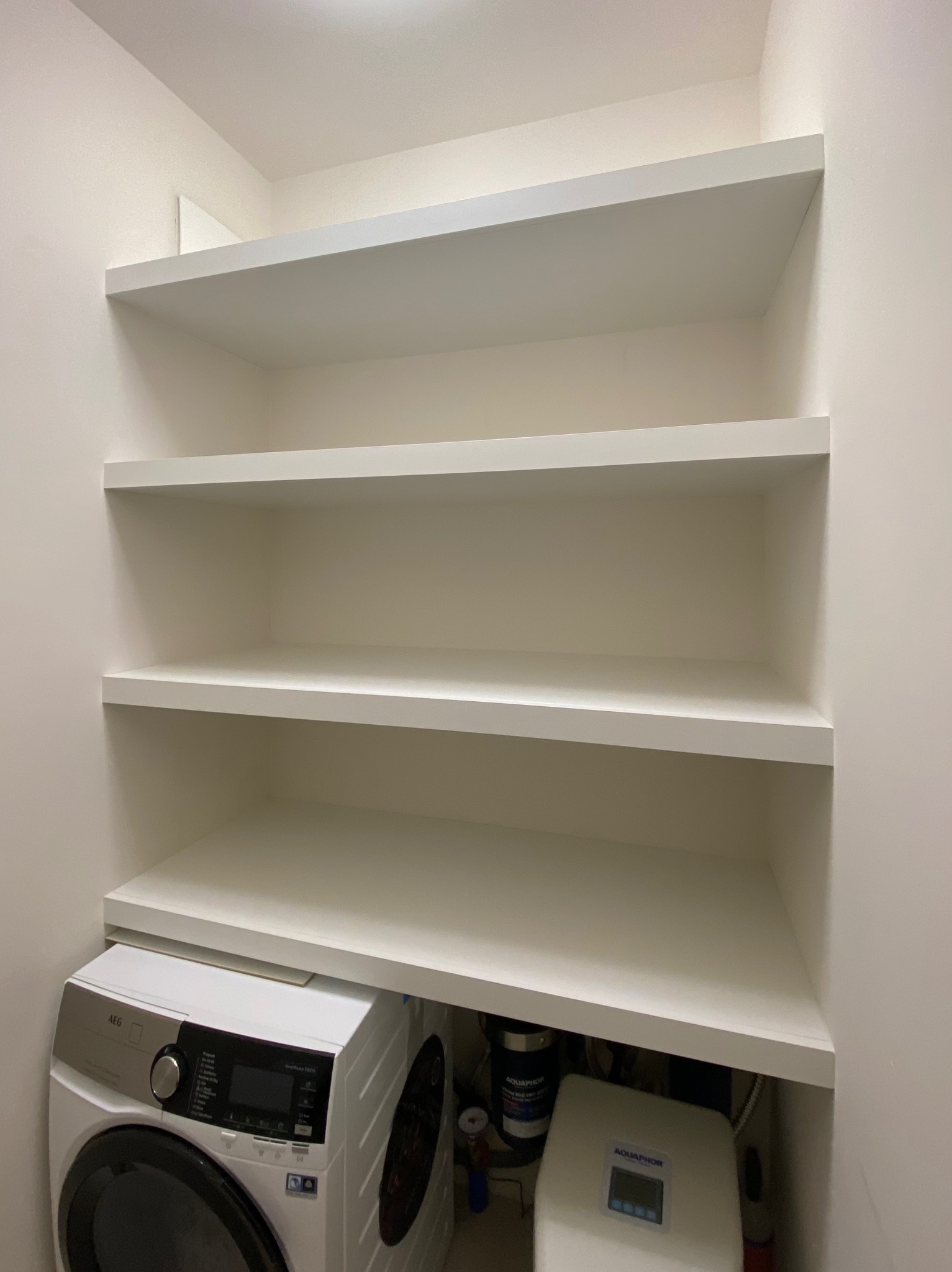 Białe, proste półki na wymiar w pomieszczeniu gospodarczym, z pralką AEG i systemem uzdatniania wody Aquaphor pod najniższą półką.