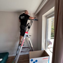 LESKI-KLIM - Mężczyzna na drabinie montuje jednostkę wewnętrzną klimatyzacji przy oknie, widoczny karton z opisem urządzenia Sinclair SOH-12BIK2.