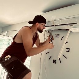 LESKI-KLIM - Mężczyzna w czapce i okularach instaluje klimatyzator ścienny, używając narzędzi; widoczny fragment zegara na ścianie.