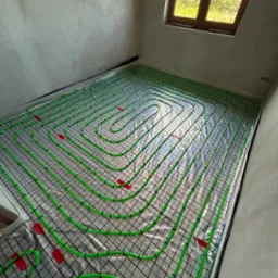 Instalacja ogrzewania podłogowego: zielone rurki ułożone spiralnie na czarnej siatce, przymocowane czerwonymi klipsami, widok z góry w surowym wnętrzu z oknem.