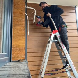 LESKI-KLIM - Uśmiechnięty monter na drabinie, instaluje przewody klimatyzacji na elewacji budynku z drewnopodobną okładziną, widoczny wiertarko-wkrętak Bosch.