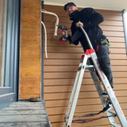 Uśmiechnięty monter na drabinie, instaluje przewody klimatyzacji na elewacji budynku z drewnopodobną okładziną, widoczny wiertarko-wkrętak Bosch.