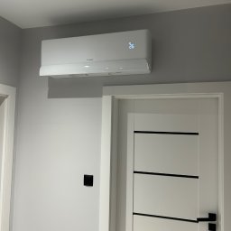 LESKI-KLIM - Biały klimatyzator z wyświetlaczem temperatury (26°C) zamontowany na szarej ścianie nad białą, nowoczesną, wewnętrzną stolarką drzwiową z czarnymi akcentami.