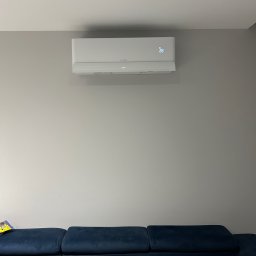 LESKI-KLIM - Biały klimatyzator ścienny z wyświetlaczem temperatury (28 stopni) zamontowany nad granatową sofą w szarym pokoju. Widoczny okrągły nawiewnik sufitowy.