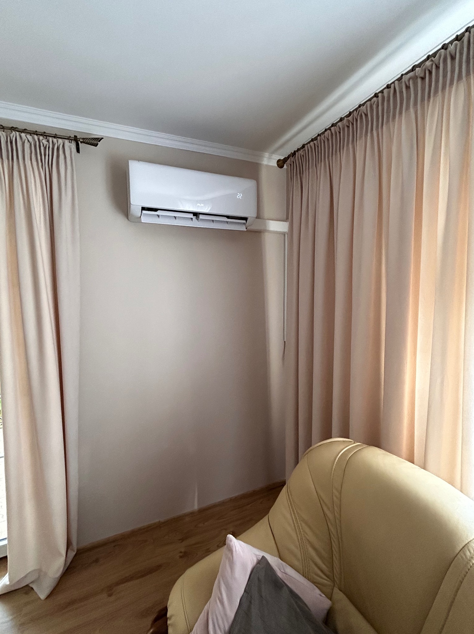 Biały klimatyzator ścienny marki AUX zamontowany w jasnym pokoju z beżowymi zasłonami i częściowo widocznym skórzanym fotelem.