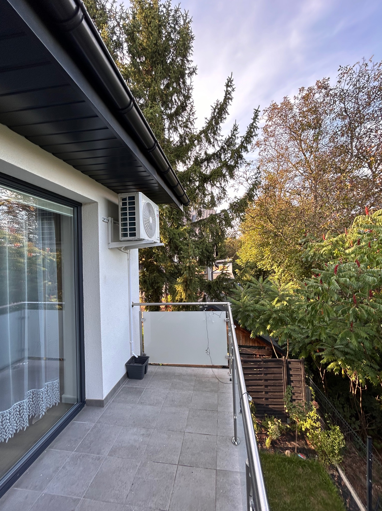 Biała jednostka zewnętrzna klimatyzacji zamontowana na ścianie budynku, widok z balkonu z szarymi płytkami i metalową balustradą, na tle zieleni drzew i krzewów.