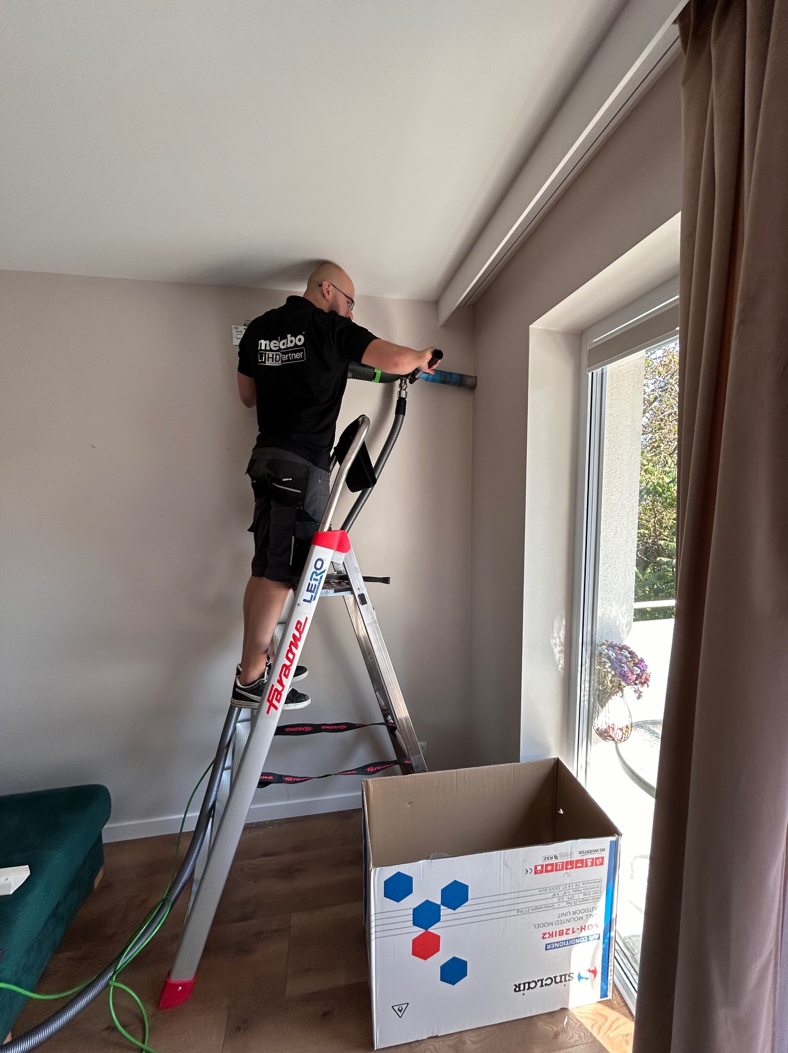Mężczyzna na drabinie montuje jednostkę wewnętrzną klimatyzacji przy oknie, widoczny karton z opisem urządzenia Sinclair SOH-12BIK2.