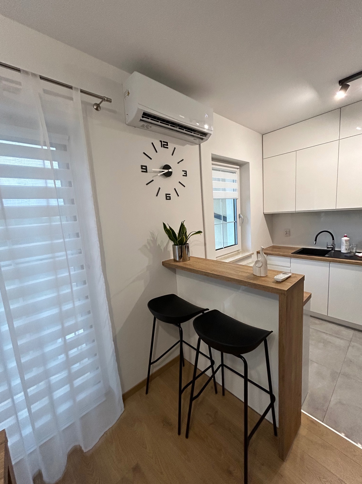 Nowoczesna kuchnia z klimatyzatorem, zegarem ściennym i dwoma czarnymi hokerami przy drewnianym blacie.