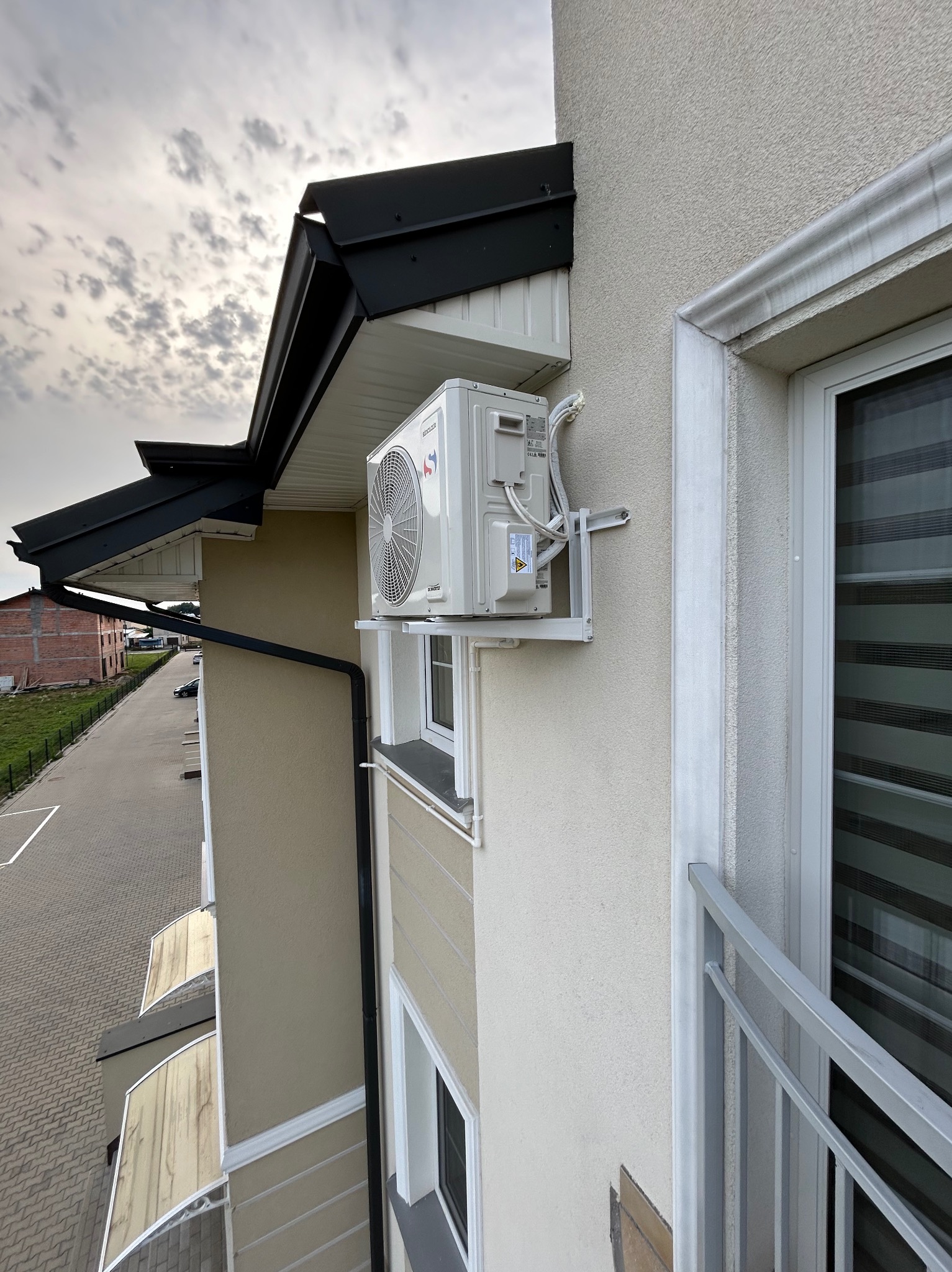 Biała jednostka zewnętrzna klimatyzacji zamontowana na ścianie budynku z beżową elewacją, widok z góry na parking i budynek w budowie w tle.