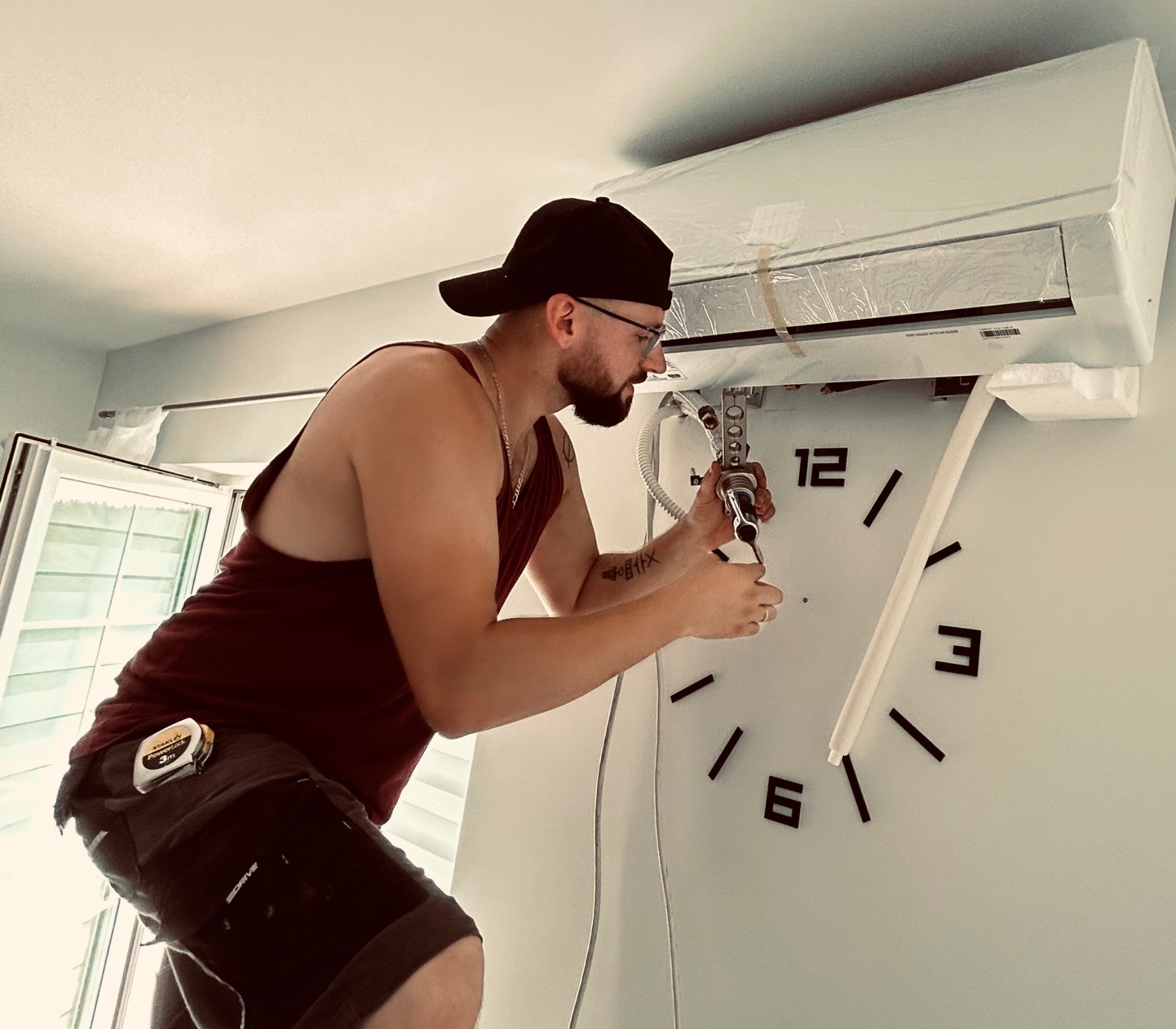 Mężczyzna w czapce i okularach instaluje klimatyzator ścienny, używając narzędzi; widoczny fragment zegara na ścianie.