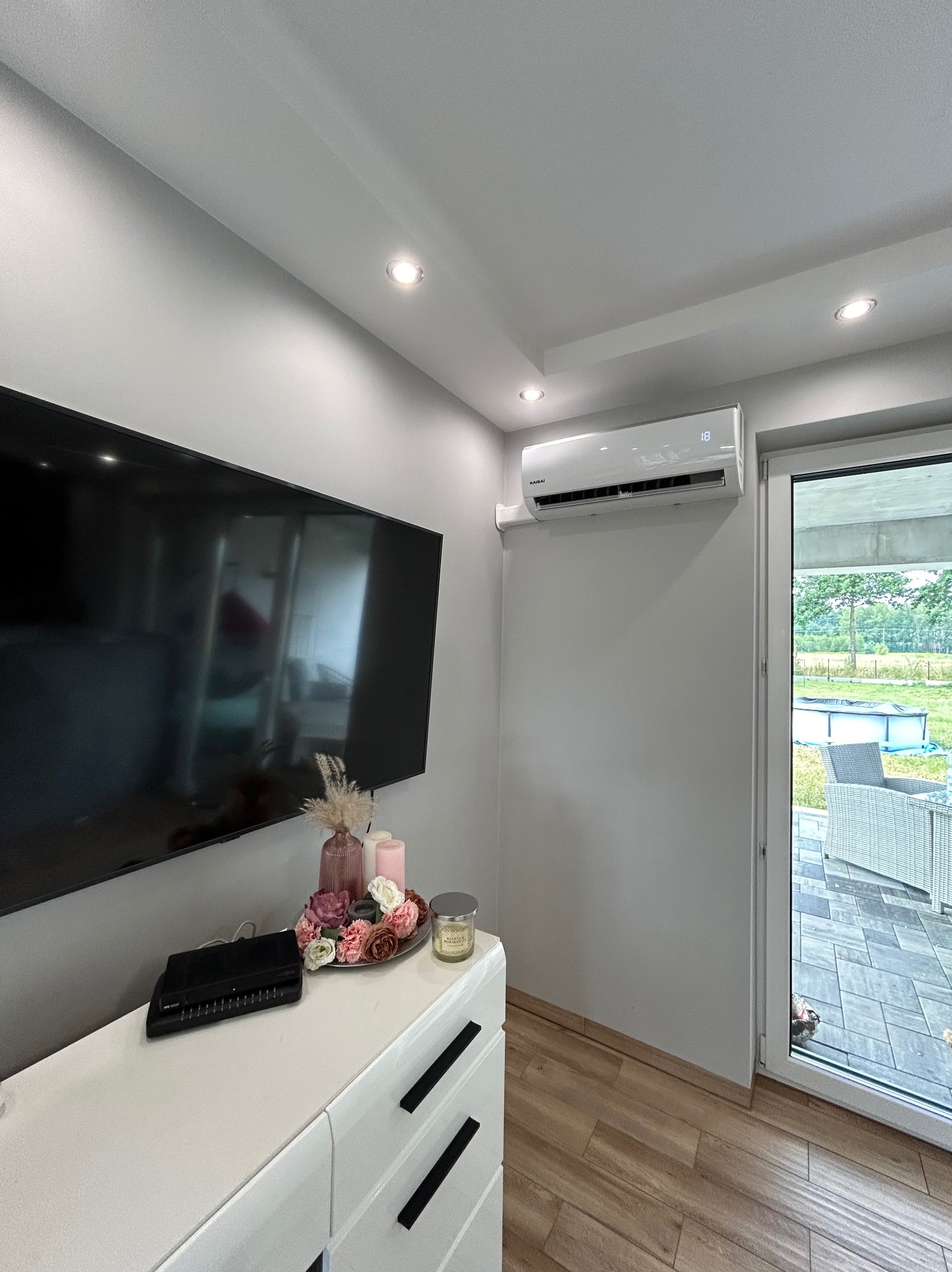 Nowoczesny klimatyzator Kaisai zamontowany w salonie nad drzwiami balkonowymi, widok na taras z basenem, dekoracje na komodzie pod telewizorem.