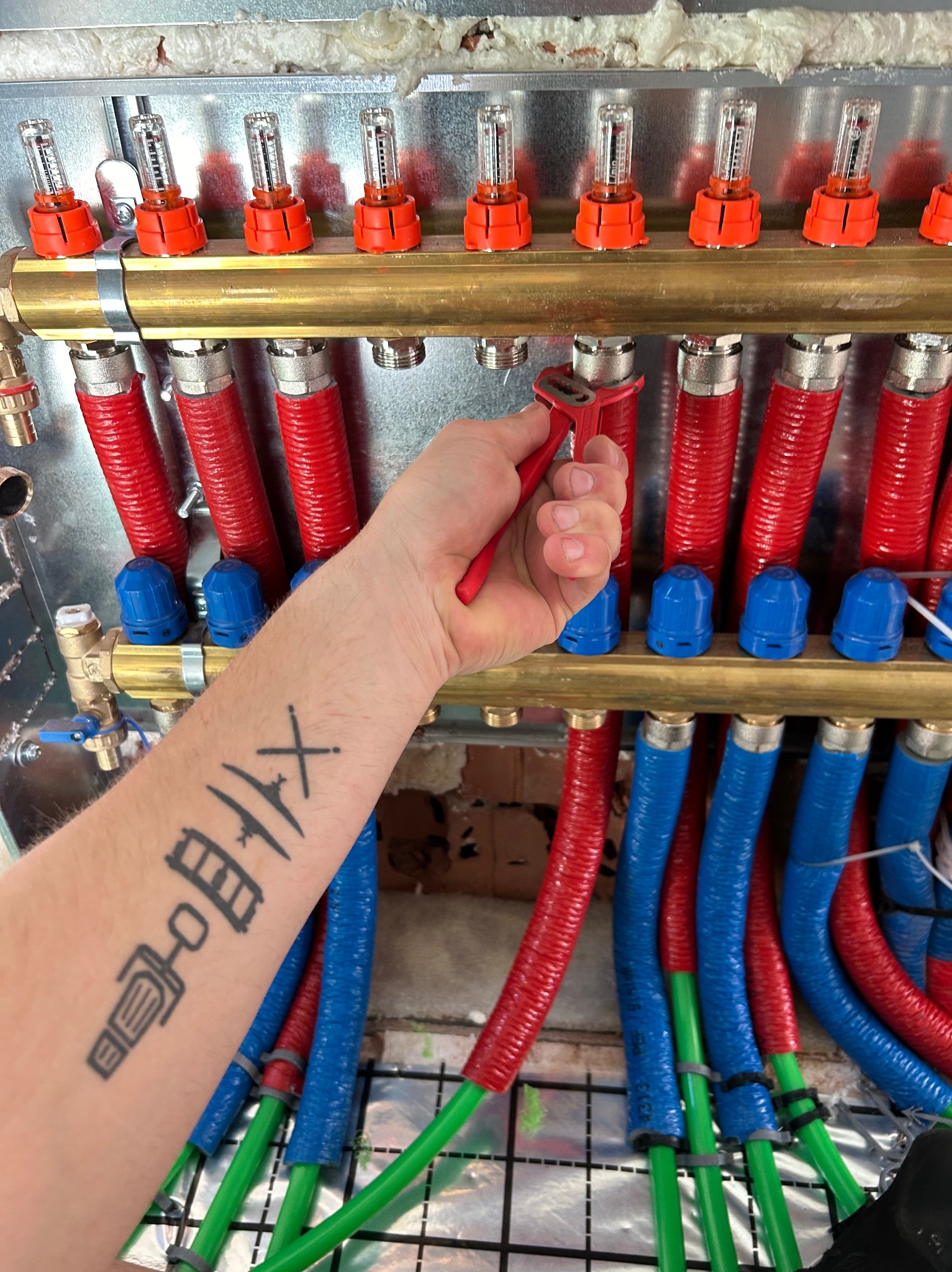 Instalator z tatuażem na ramieniu reguluje czerwonym kluczem rozdzielacz ogrzewania podłogowego z czerwono-niebieskimi rurami.