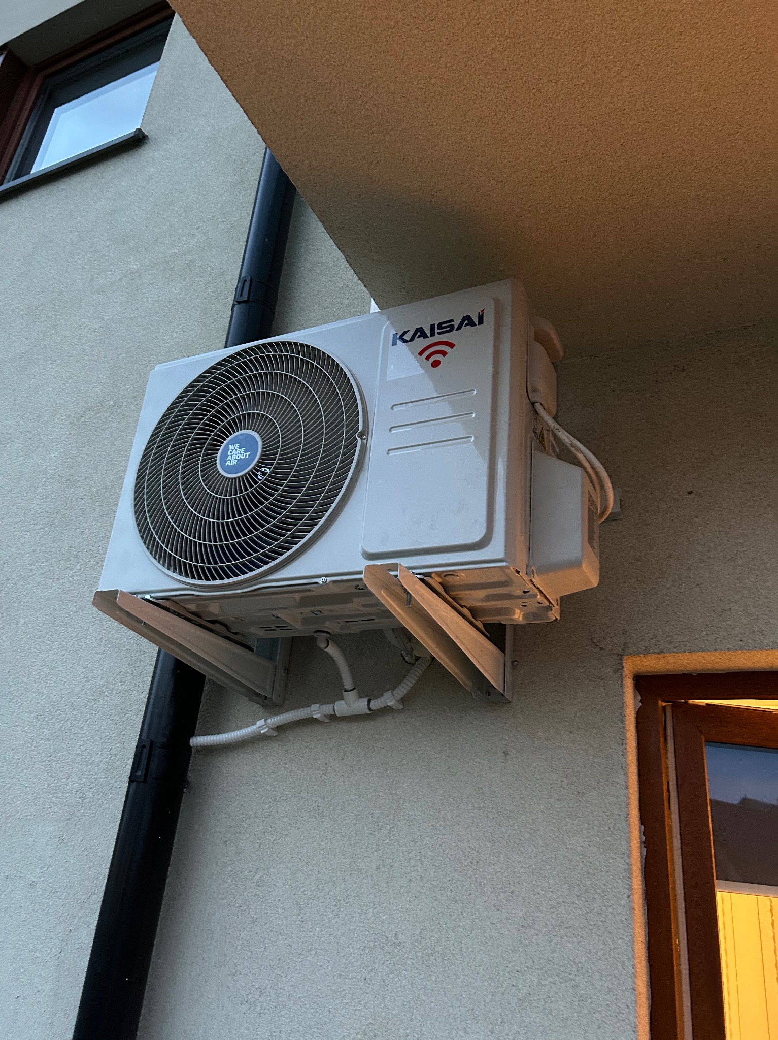 Zewnętrzna jednostka klimatyzacji Kaisai zamontowana na ścianie budynku, widoczne rury odprowadzające skropliny i przewody elektryczne, obok okno i rura spustowa.