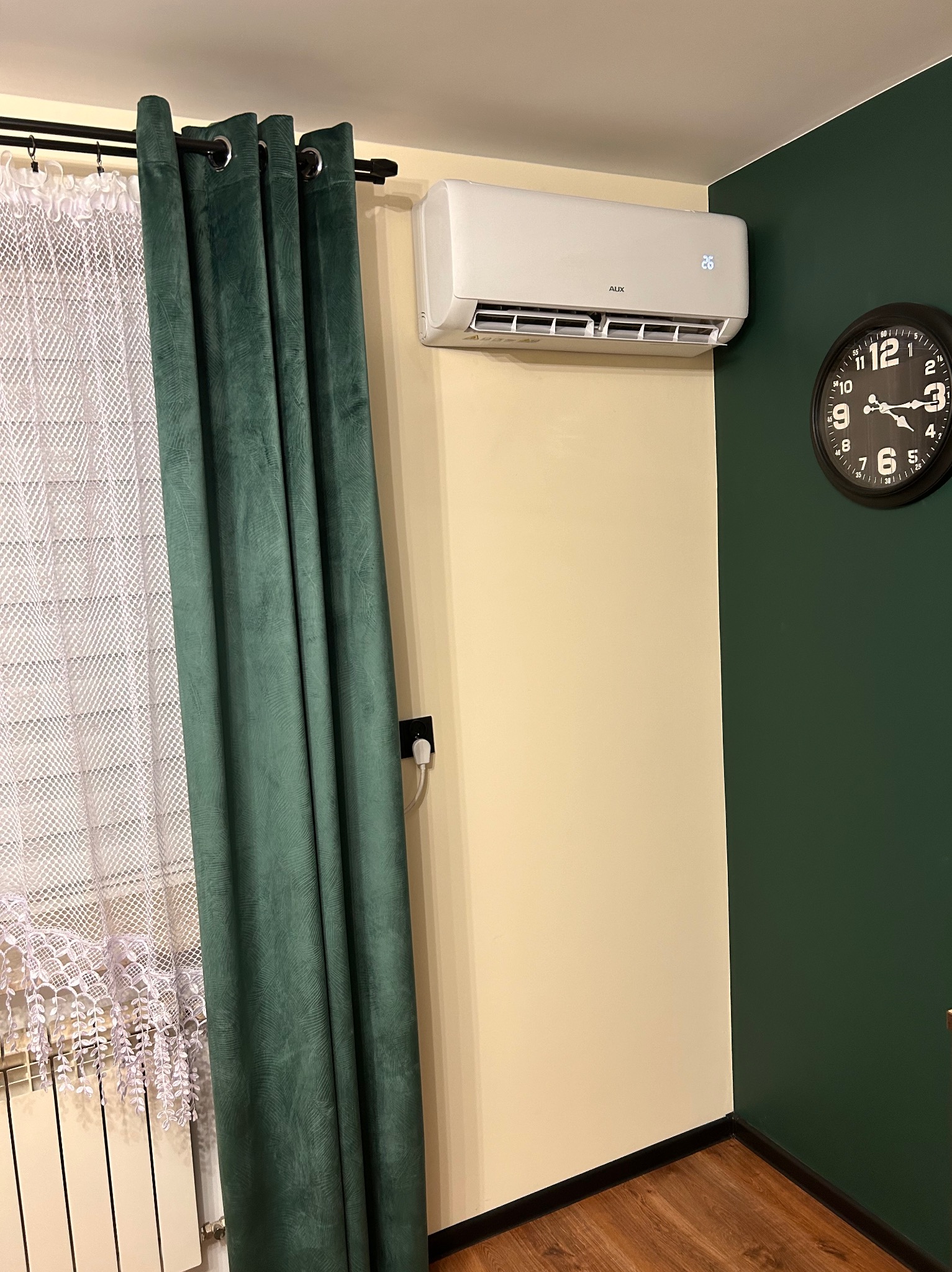 Biały klimatyzator ścienny marki AUX z wyświetlaczem temperatury, zamontowany w pomieszczeniu z beżową ścianą i zielonym akcentem, obok wiszący czarny zegar, zasłonięte okno z grzejnikiem pod spodem.