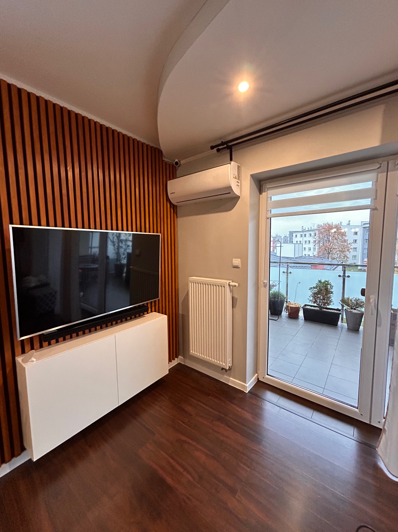 Nowoczesny salon z zamontowaną klimatyzacją, telewizorem na drewnianej ścianie i wyjściem na balkon z roślinami w donicach.
