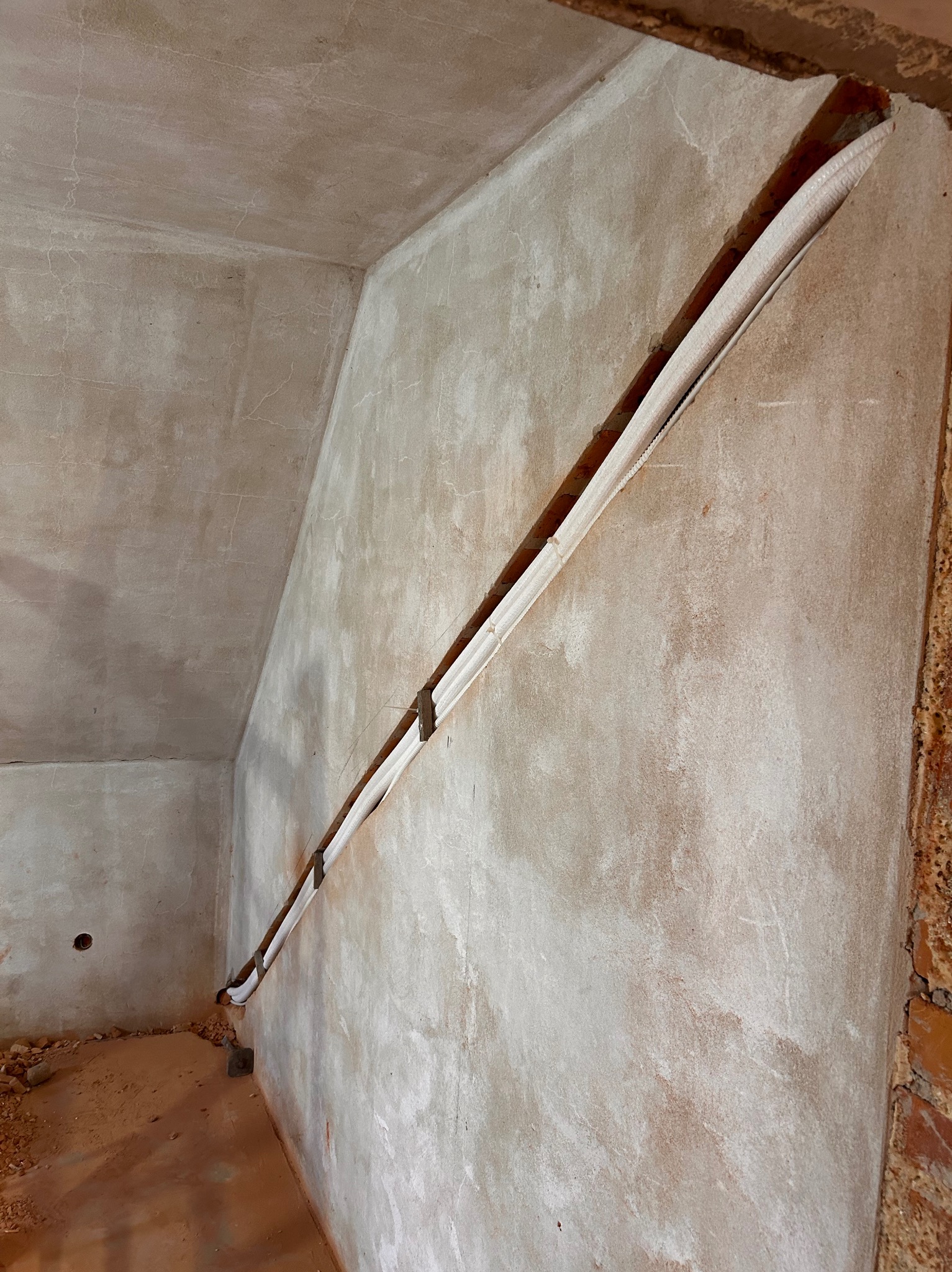 Wnętrze w trakcie remontu: białe rury instalacyjne prowadzone ukośnie w bruździe wyciętej w surowej, szarej ścianie z widocznymi śladami zaprawy i gruzem na podłodze.