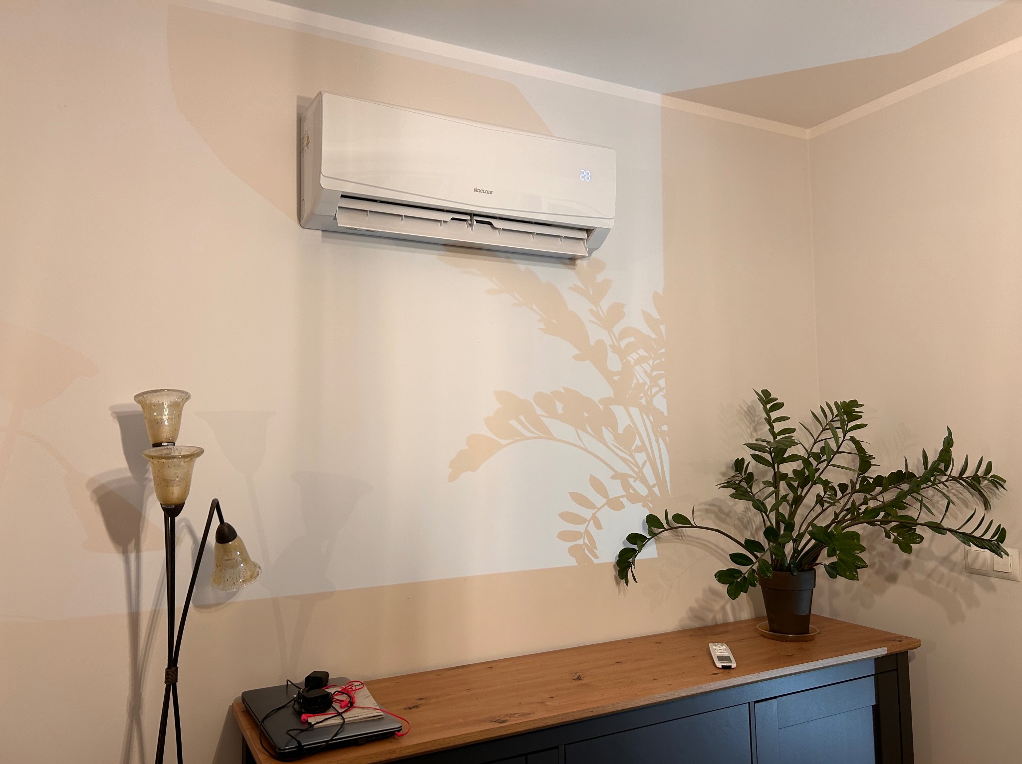 Biały klimatyzator Sinclair zamontowany na beżowej ścianie w pokoju, widoczny wyświetlacz temperatury 28 stopni, cień rośliny doniczkowej na ścianie, obok lampa stojąca i komoda.