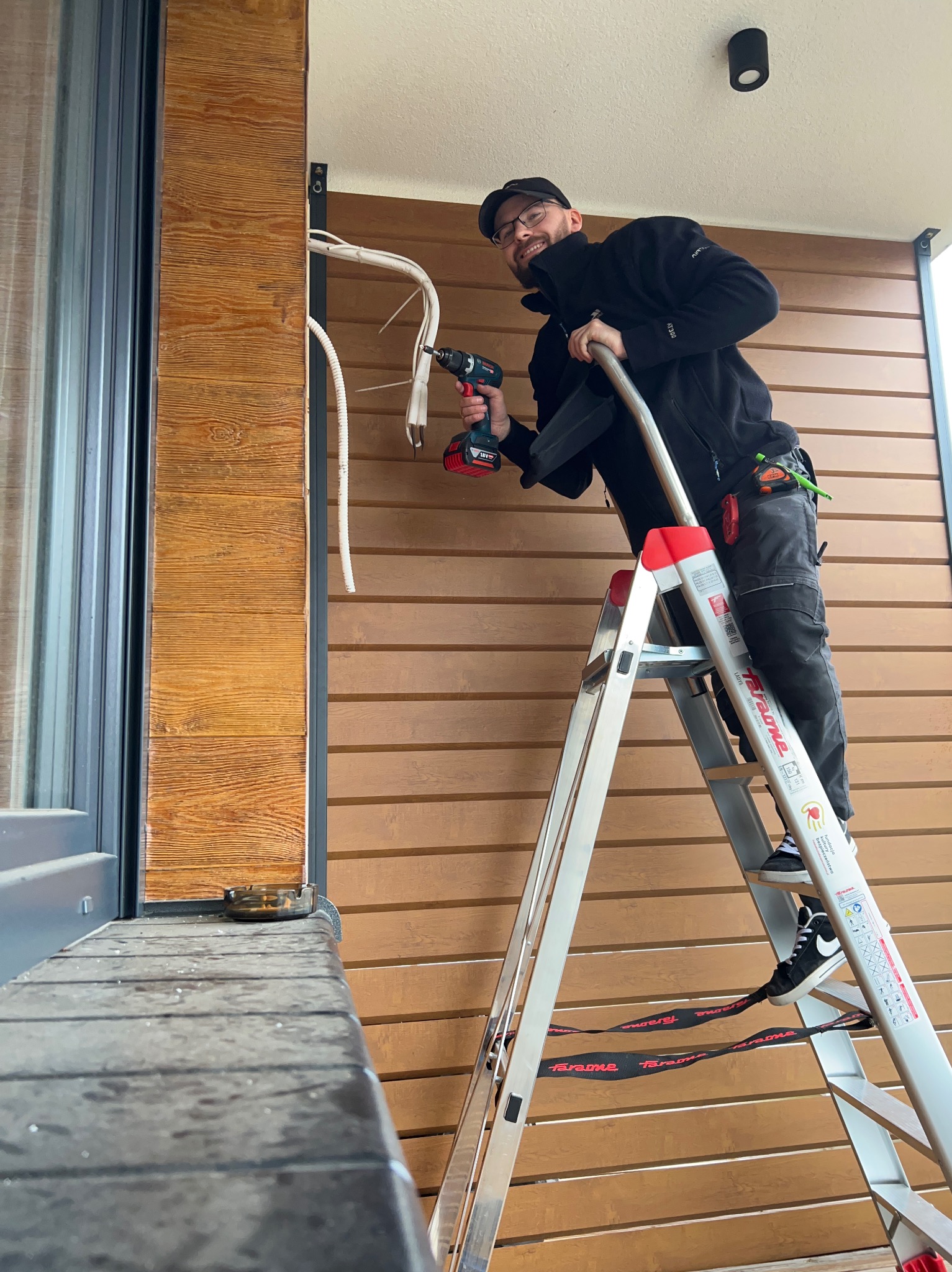 Uśmiechnięty monter na drabinie, instaluje przewody klimatyzacji na elewacji budynku z drewnopodobną okładziną, widoczny wiertarko-wkrętak Bosch.