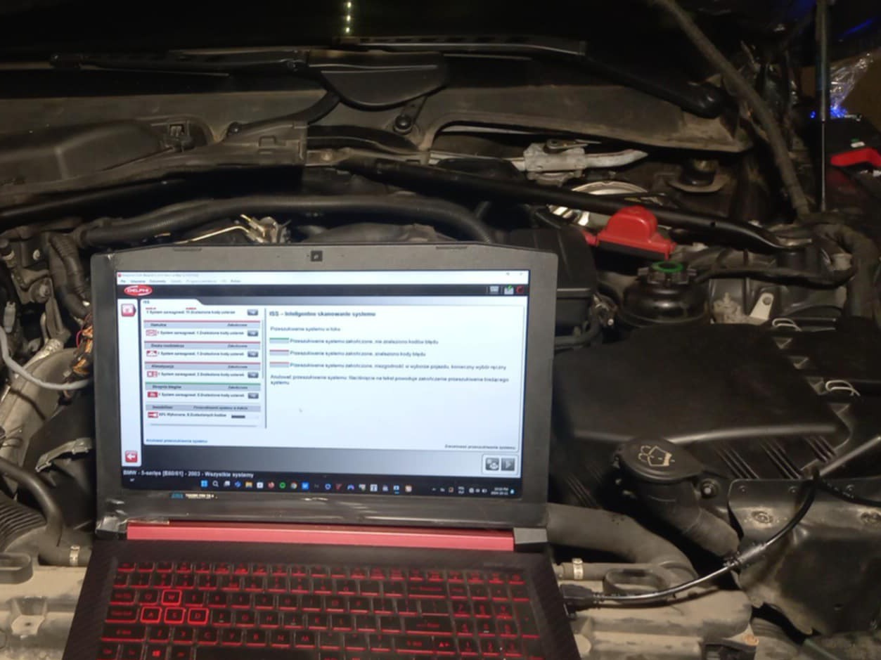 Laptop z uruchomionym oprogramowaniem diagnostycznym podłączony do silnika BMW serii 5 (E60/E61) z 2003 roku, widok na otwartą maskę i elementy silnika.