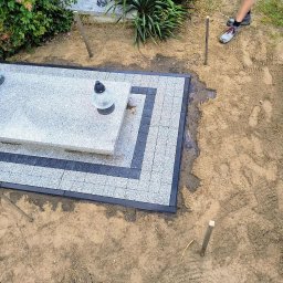 Błażej Łabędzki - Wykończony nagrobek z jasnoszarą kostką i ciemnoszarym obramowaniem. Widoczne kwiaty, znicze i drewniane kołki w ziemi. Ujęcie z góry.