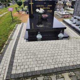 Błażej Łabędzki - Czarny nagrobek z krzyżem i kwiatami na cmentarzu, otoczony jasną kostką brukową z ciemnymi pasami. W tle inne groby i zieleń.