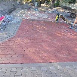KBM Bruk - Częściowo ułożona, czerwona kostka brukowa na podjeździe, z widocznym przygotowanym podłożem i narzędziami. Fragment szarej kostki na dole kadru.