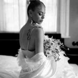 My Little White Studio - Czarno-białe zdjęcie kobiety z upiętymi włosami, w satynowej bieliźnie i szlafroku z piórami, siedzącej na łóżku i trzymającej bukiet kwiatów, z widocznym tatuażem na plecach.