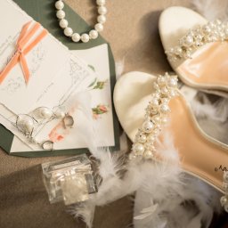 My Little White Studio - Eleganckie białe buty ślubne z perłami, zaproszenie ślubne z różową wstążką, perłowy naszyjnik, kolczyki z perłami i flakon perfum na teksturowanym tle.