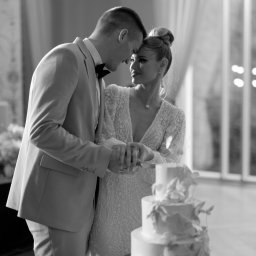 My Little White Studio - Czarno-białe zdjęcie pary młodej krojącej tort weselny; on w jasnym garniturze i muszce, ona w koronkowej sukni z dekoltem, oboje z czułością patrzą na siebie