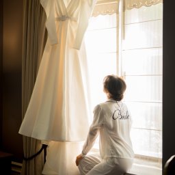 My Little White Studio - Panna młoda w białej piżamie z napisem 'Bride' na plecach, siedząca na parapecie i patrząca na wiszącą suknię ślubną przy oknie z dekoracyjnymi zasłonami.