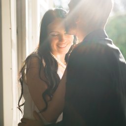 My Little White Studio - Uśmiechnięta kobieta w białej sukience i perłach, obejmowana przez mężczyznę w garniturze, oświetlona promieniami słońca wpadającymi przez okno.