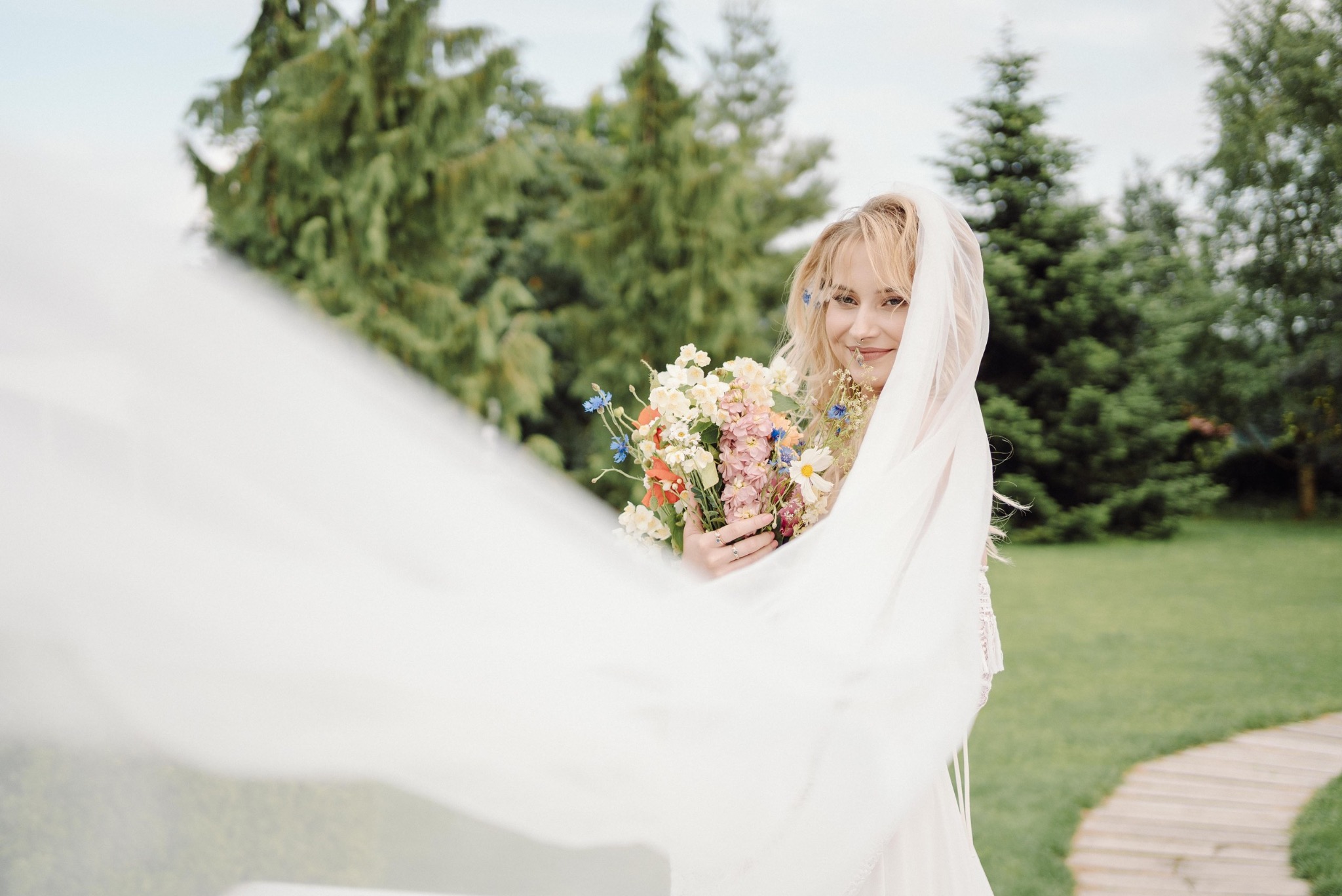 Uśmiechnięta blondynka w zwiewnym welonie, trzymająca bukiet polnych kwiatów z niebieskimi akcentami, na tle zieleni ogrodu i ścieżki z drewnianych płyt.