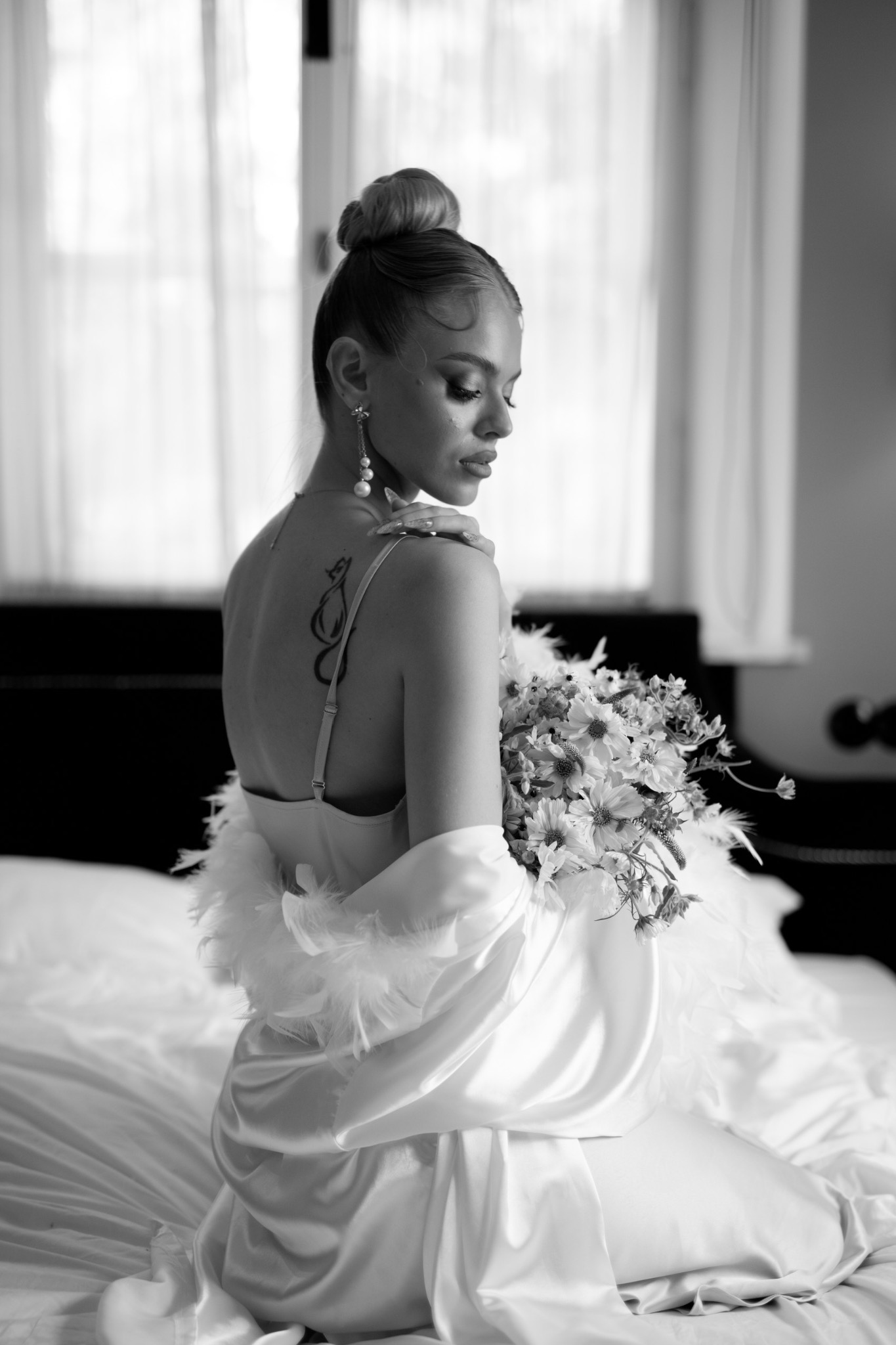 Czarno-białe zdjęcie kobiety z upiętymi włosami, w satynowej bieliźnie i szlafroku z piórami, siedzącej na łóżku i trzymającej bukiet kwiatów, z widocznym tatuażem na plecach.