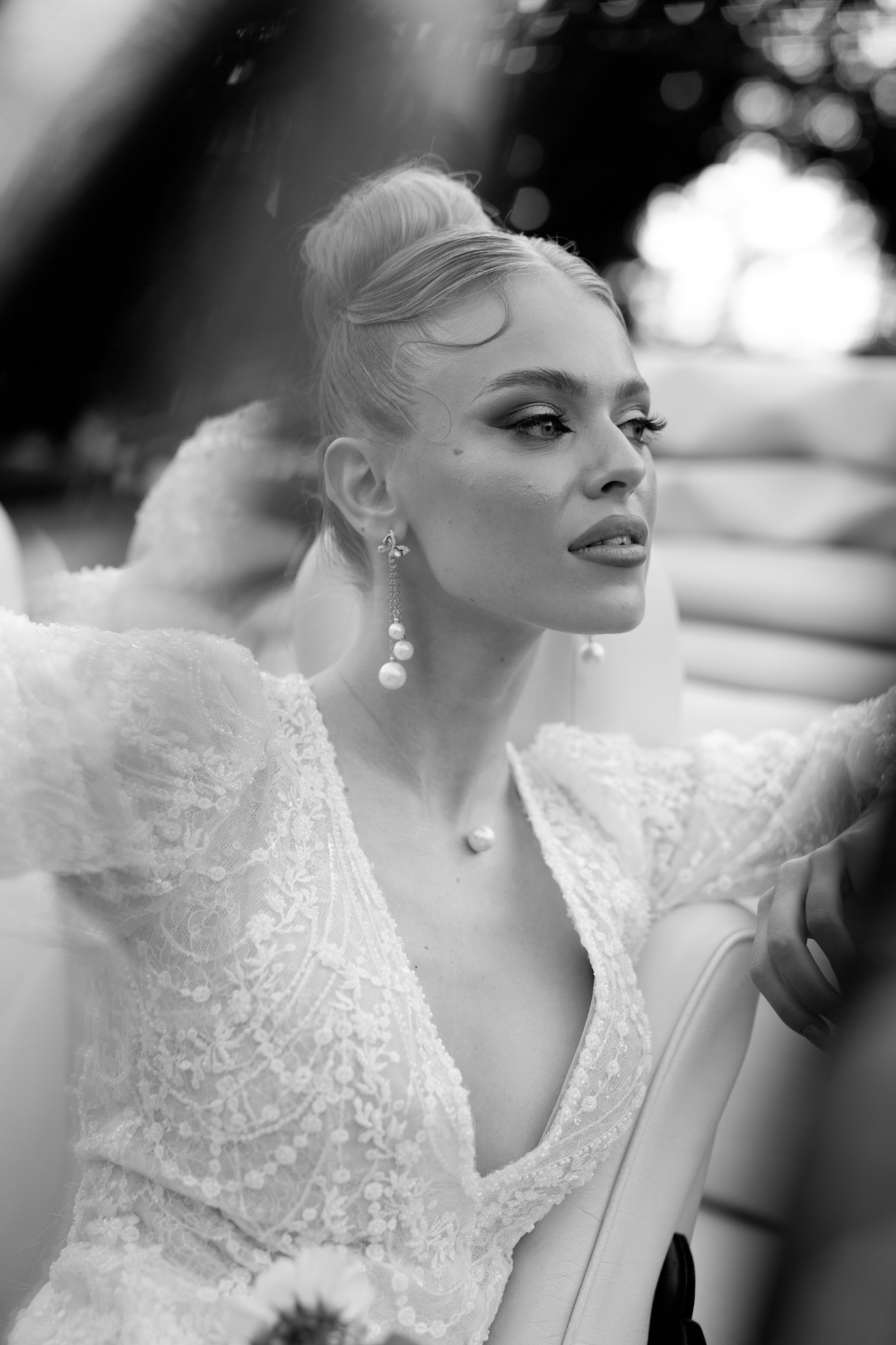 Czarno-białe, portretowe ujęcie kobiety w bogato zdobionej, koronkowej sukni z głębokim dekoltem, z perłowymi kolczykami i naszyjnikiem, włosy upięte w kok, wzrok skierowany w prawo, siedzącej...