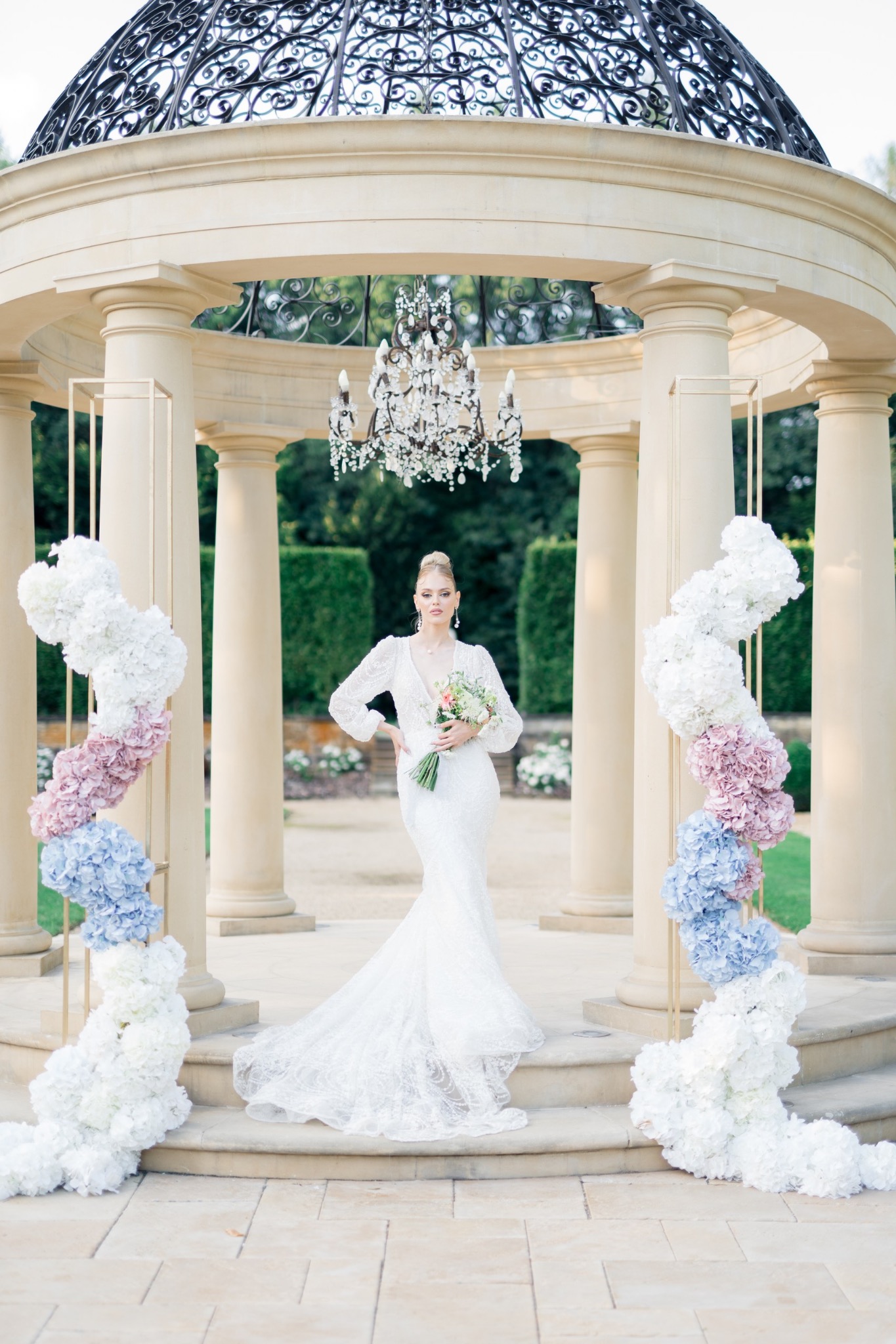 Panna młoda w koronkowej sukni pozuje z bukietem w altanie z kolumnami, ozdobionej kwiatowymi girlandami i kryształowym żyrandolem.
