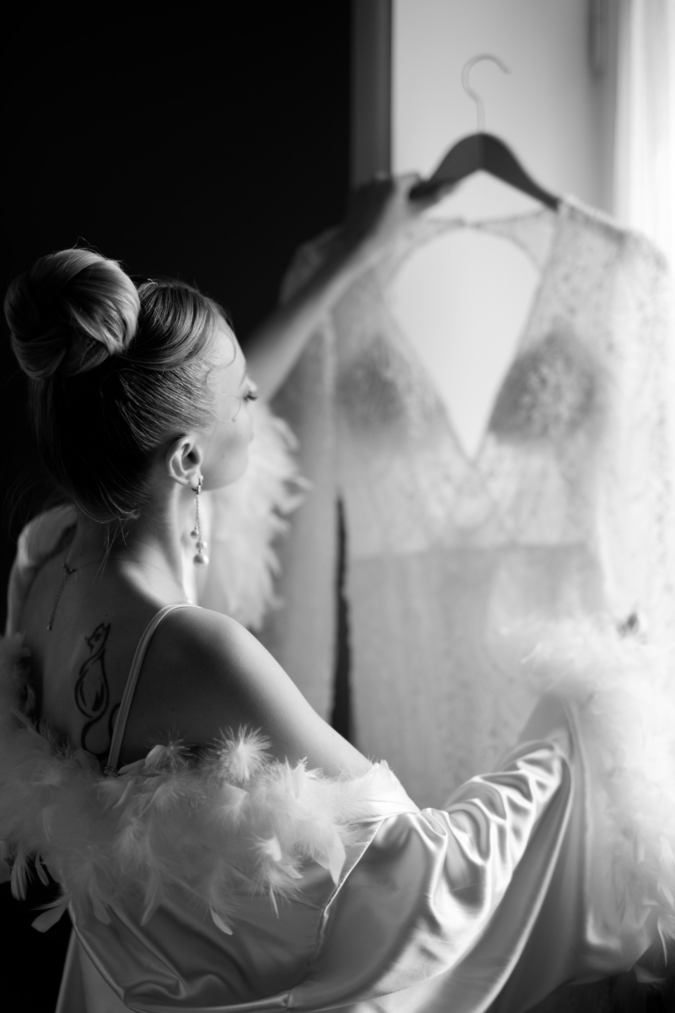 Kobieta w szlafroku z piórami ogląda koronkową sukienkę na wieszaku, widoczny tatuaż na plecach i kolczyki z perłami, zdjęcie czarno-białe.