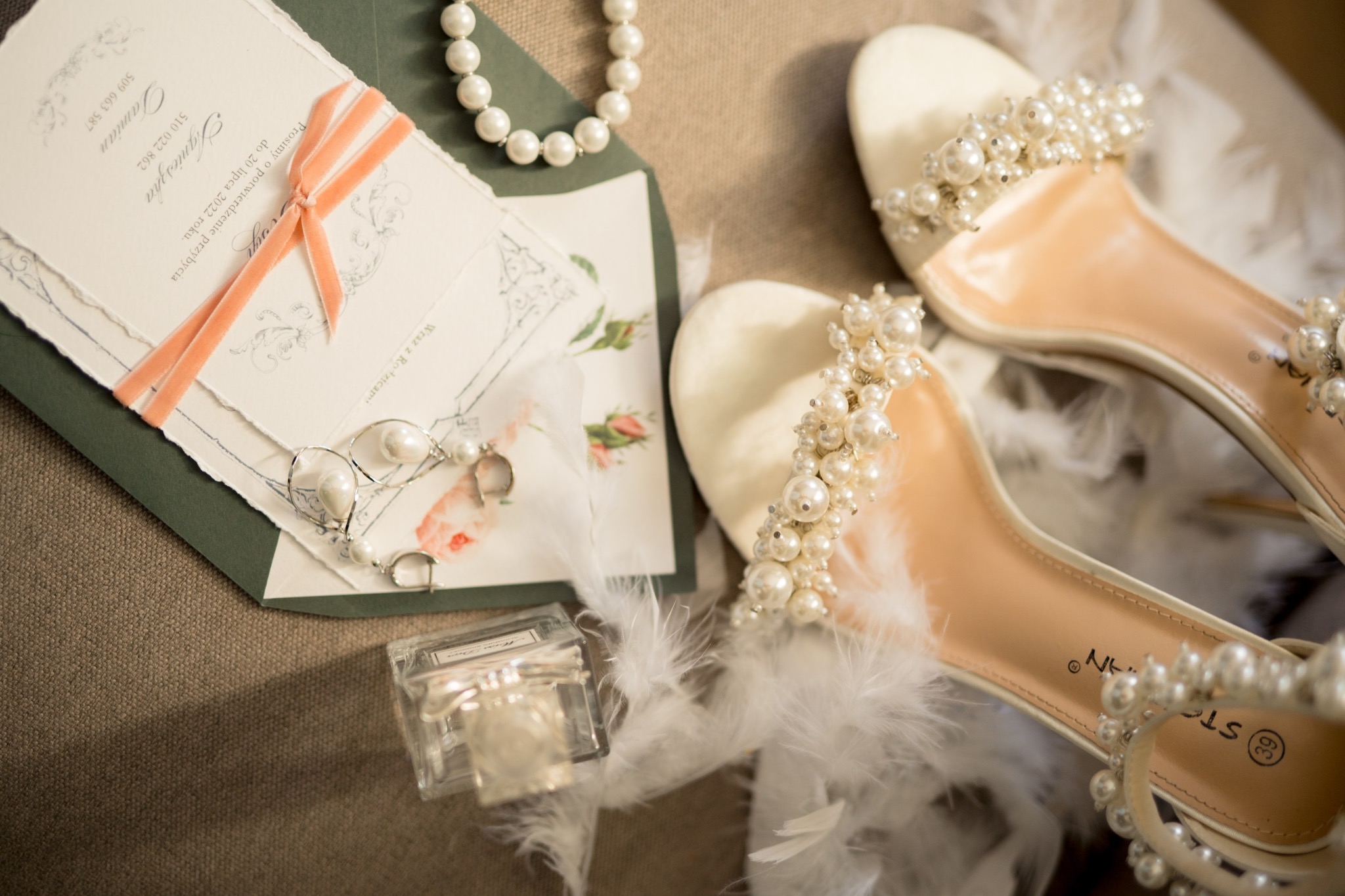 Eleganckie białe buty ślubne z perłami, zaproszenie ślubne z różową wstążką, perłowy naszyjnik, kolczyki z perłami i flakon perfum na teksturowanym tle.