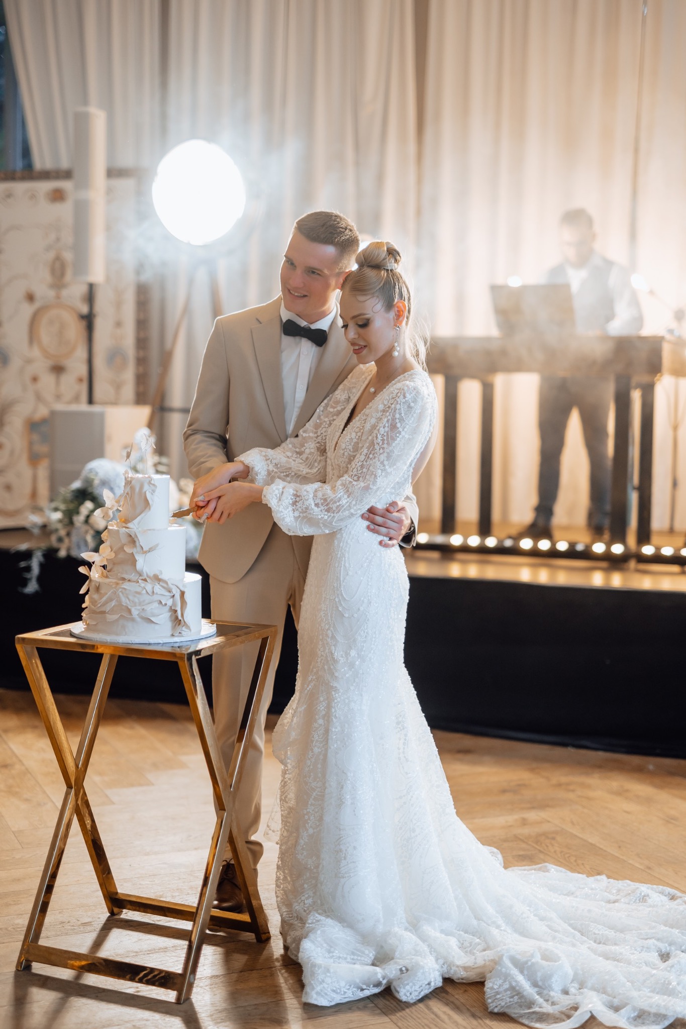 Para młoda kroi trzypiętrowy tort weselny na złotym stoliku, w tle DJ na podwyższeniu i dekoracje.