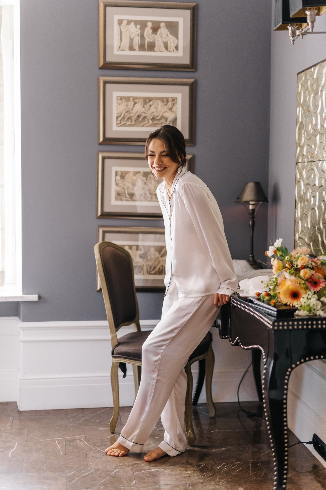 Uśmiechnięta kobieta w satynowej piżamie opiera się o czarne, zdobione biurko w pokoju z klasycznymi obrazami na ścianie i bukietem kwiatów.