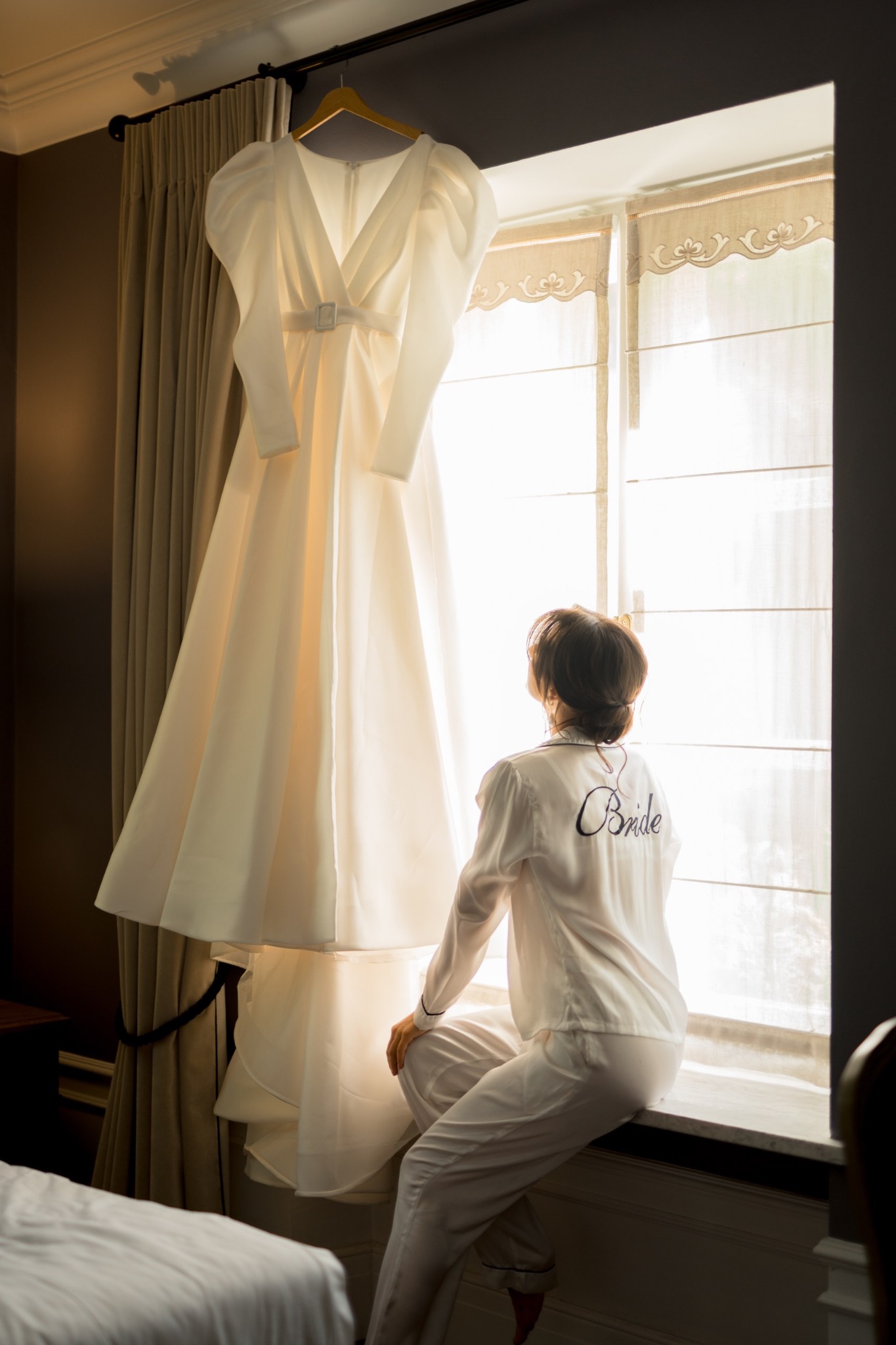 Panna młoda w białej piżamie z napisem 'Bride' na plecach, siedząca na parapecie i patrząca na wiszącą suknię ślubną przy oknie z dekoracyjnymi zasłonami.