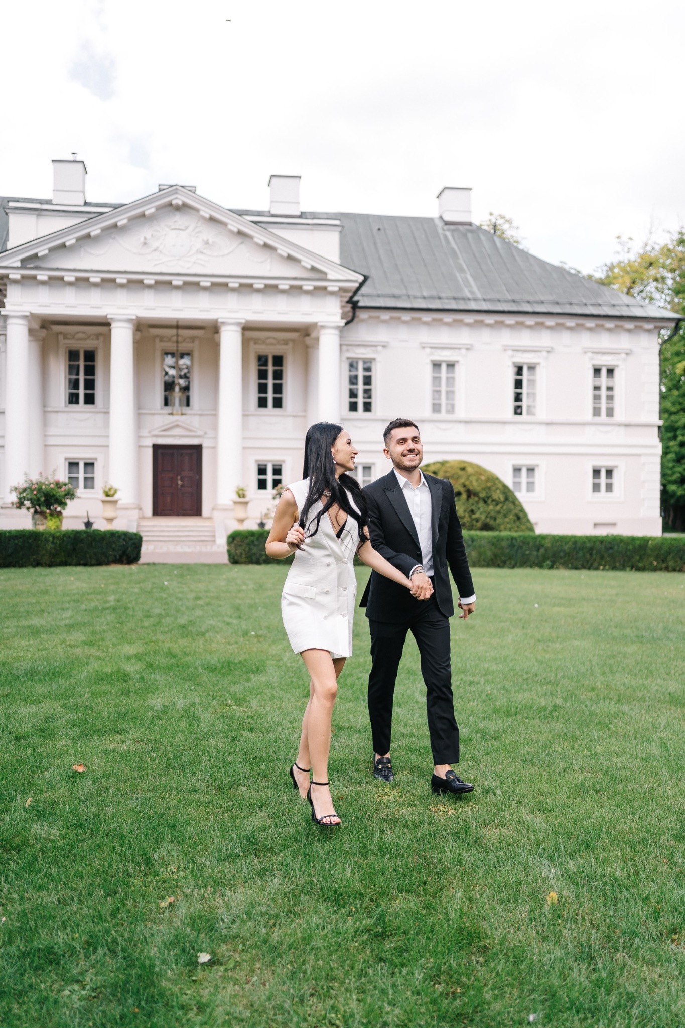Uśmiechnięta para, kobieta w białej sukience i mężczyzna w czarnym garniturze, spacerują trzymając się za ręce na tle klasycystycznego pałacu z kolumnami i rozległym trawnikiem.