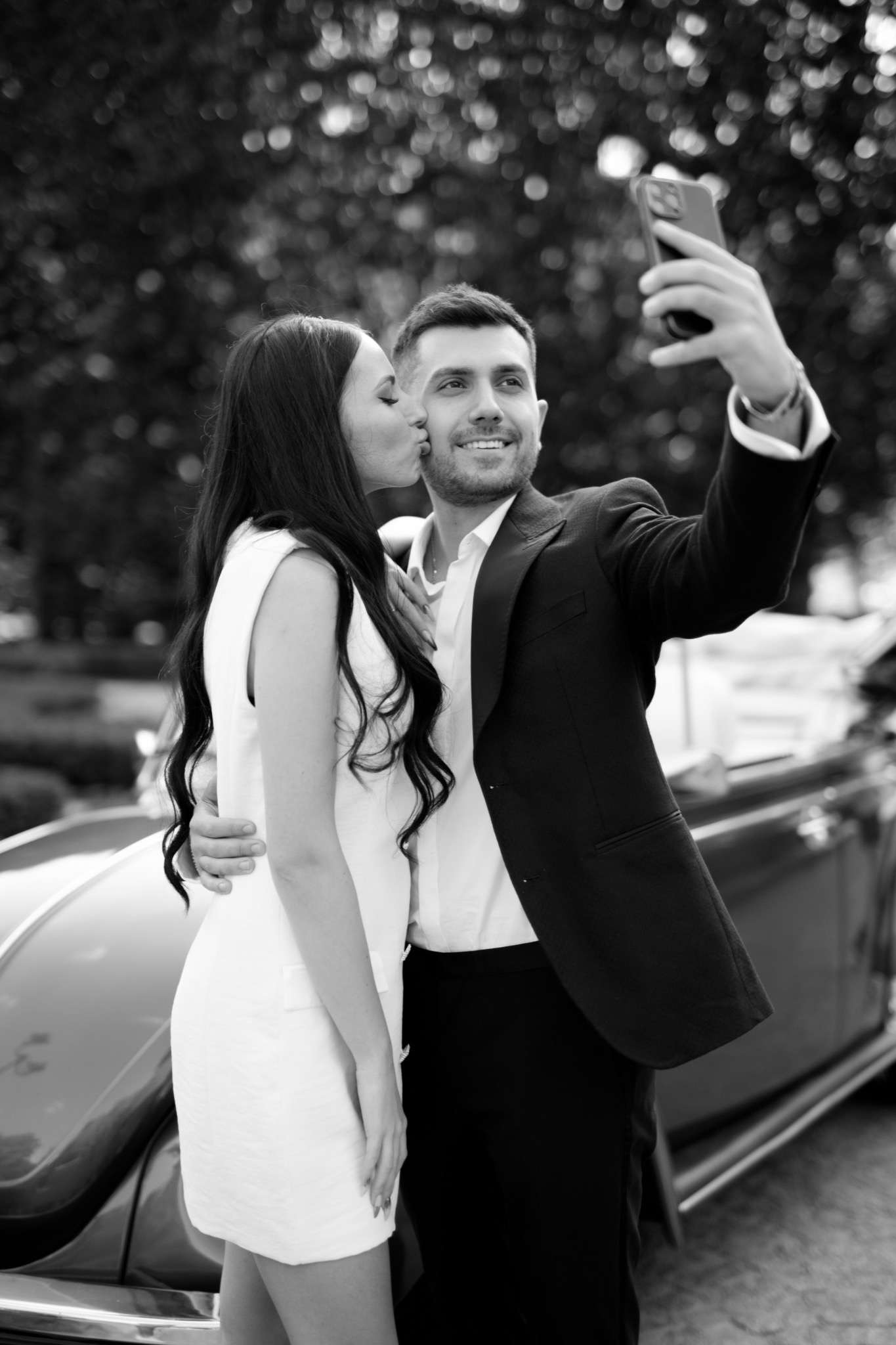 Czarno-białe zdjęcie uśmiechniętej pary robiącej selfie przed klasycznym samochodem; kobieta całuje mężczyznę w policzek.
