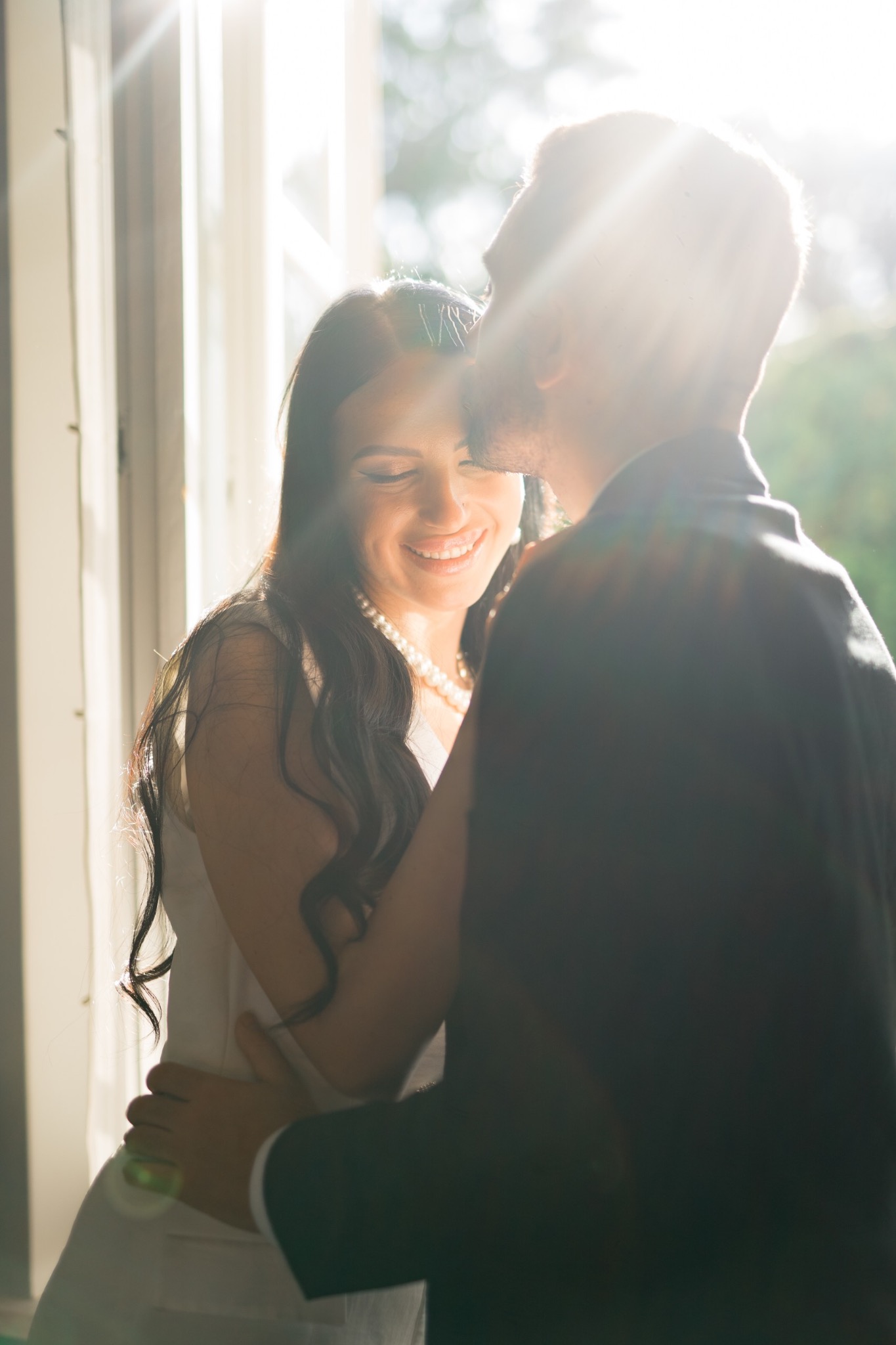 Uśmiechnięta kobieta w białej sukience i perłach, obejmowana przez mężczyznę w garniturze, oświetlona promieniami słońca wpadającymi przez okno.