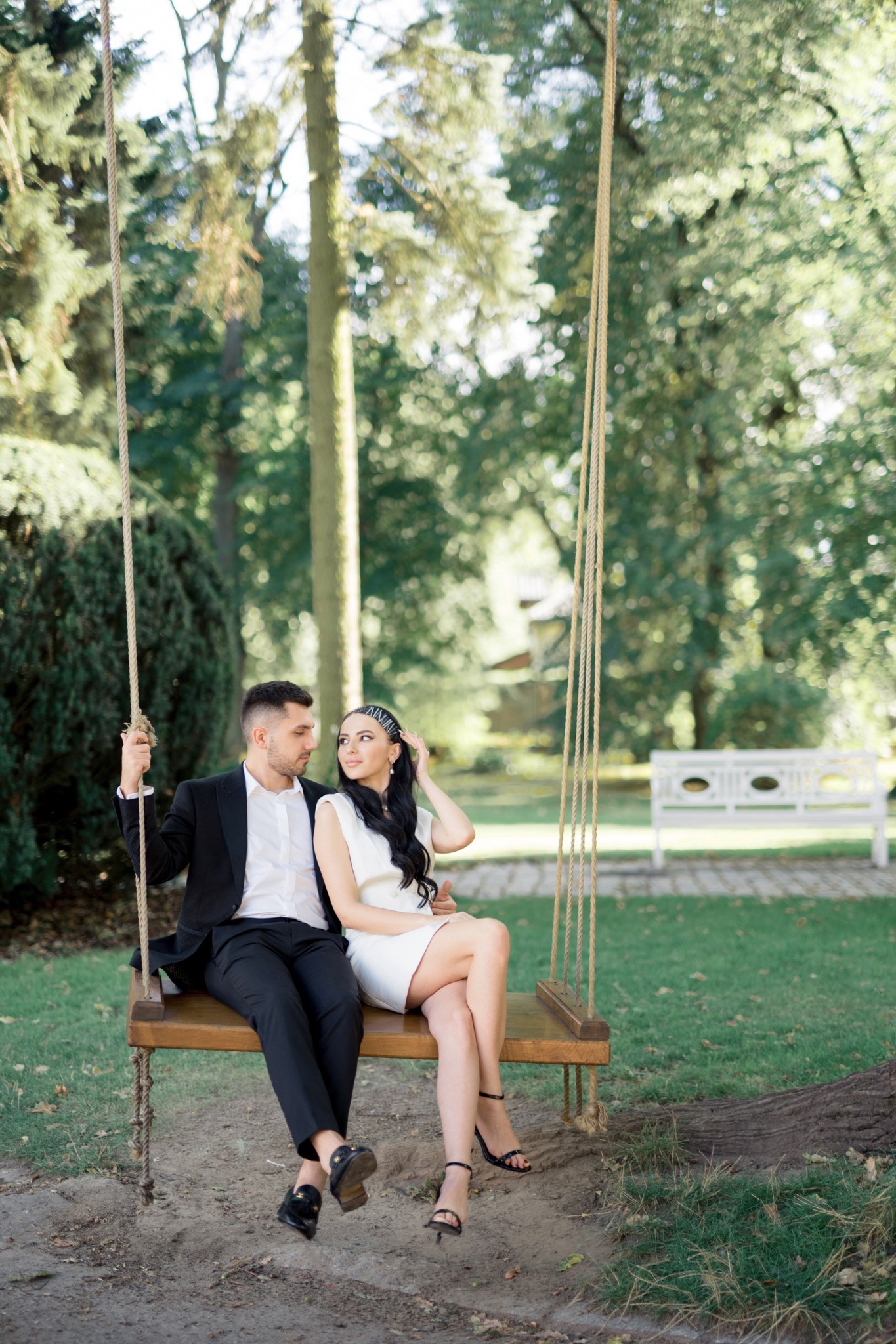 Elegancka para, mężczyzna w czarnym garniturze i kobieta w białej sukience, siedzą na drewnianej huśtawce zawieszonej na linach w parku, patrząc na siebie z uśmiechem.
