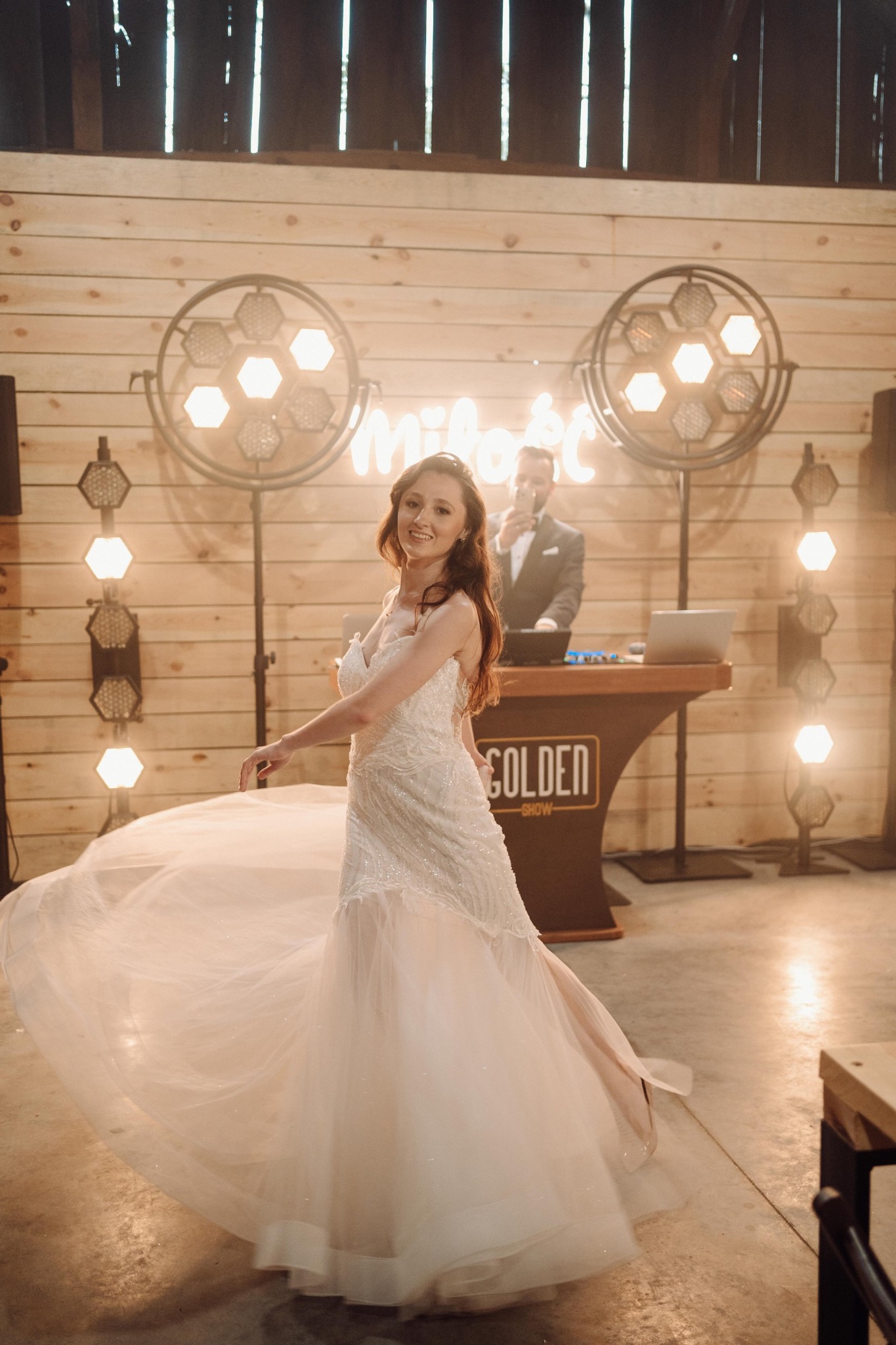 Uśmiechnięta panna młoda w sukni wiruje na parkiecie, w tle DJ z laptopem i podświetlany napis 'Miłość' oraz dekoracyjne lampy.