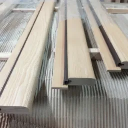 Elementy drewniane, prawdopodobnie ościeżnice lub ramiaki, z widocznym profilem i uszczelką, ułożone na tekturze falistej.