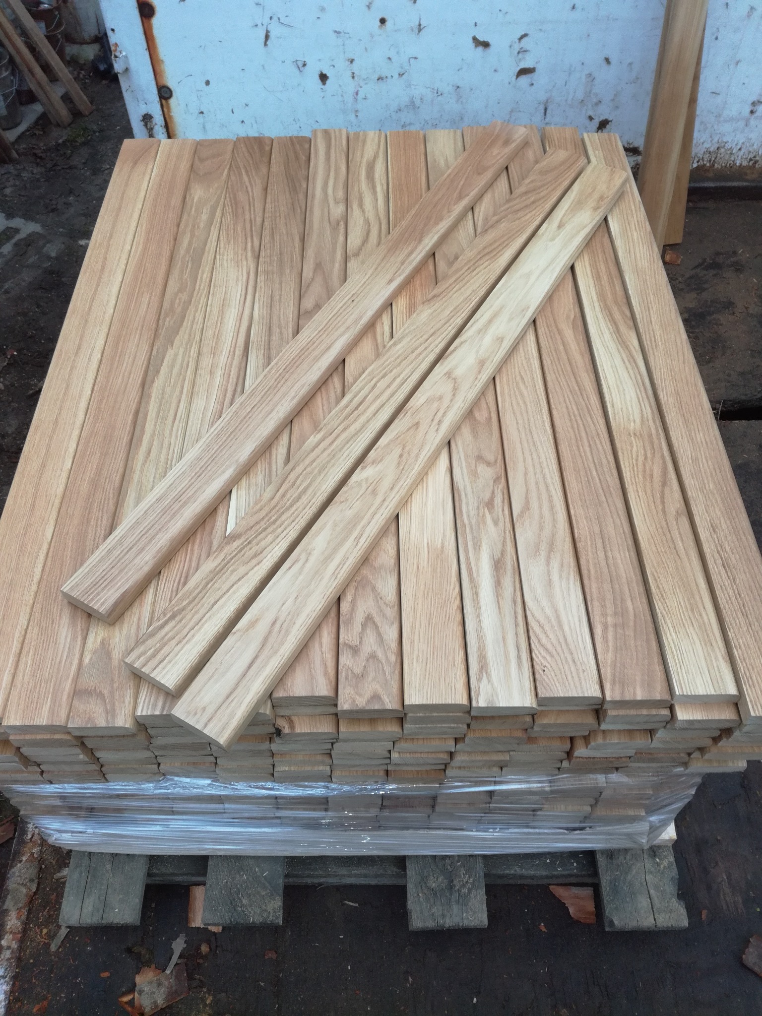 Stos surowych, jasnych listew drewnianych, zabezpieczonych folią, ułożonych na palecie, widoczne słoje drewna.